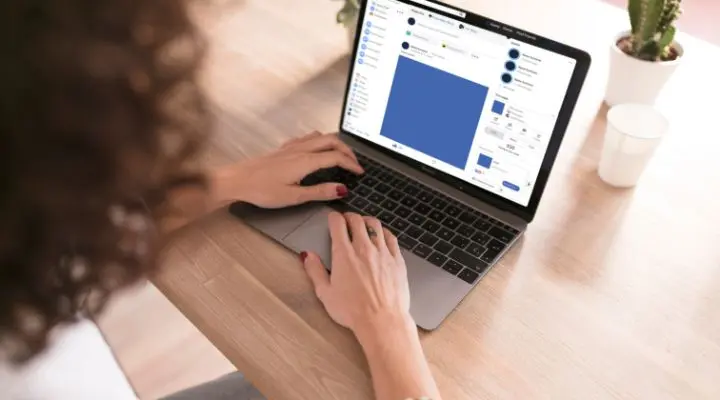 Persona que usa una computadora portátil en un escritorio de madera, ajustando la configuración de diseño gráfico en la pantalla para seguir la regla 20 % de texto en Facebook, con un cactus en maceta y una taza cerca.