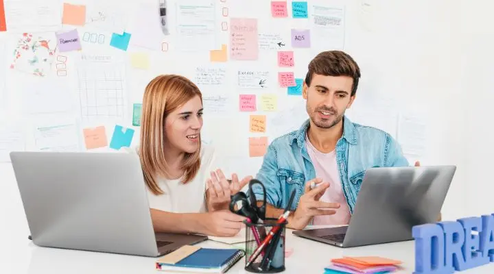 Dos personas se sientan en un escritorio con computadoras portátiles, discutiendo la importancia del Endomarketing y gesticulando, frente a una pared cubierta de notas y gráficos coloridos.