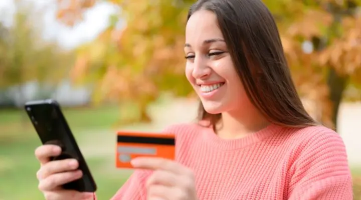 Una mujer con un suéter rosa sostiene un teléfono inteligente en una mano y cupones en la otra, sonriendo al aire libre con follaje otoñal de fondo porqaue ya merito