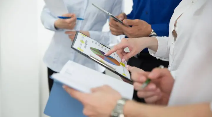 Cuatro personas sosteniendo documentos y una tableta, revisando un grÃ¡fico circular basado en encuestas pÃºblicas y tomando notas durante una reuniÃ³n o discusiÃ³n.