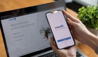 Una persona sostiene un teléfono inteligente que muestra la pantalla de inicio de sesión de LinkedIn frente a una computadora portátil que muestra la página de inicio de LinkedIn, lista para usar estrategias de empleo para mejorar su búsqueda de empleo.