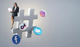 Una mujer vestida de negocios se sienta sobre un gran símbolo de hashtag, rodeada de íconos de Facebook, TikTok, Instagram y Twitter sobre un fondo gris claro, lo que muestra el poder de los hashtags efectivos para el éxito en las redes sociales.