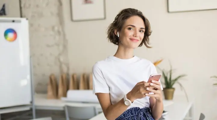 Una mujer que viste una camisa blanca y auriculares inalÃ¡mbricos sostiene un telÃ©fono inteligente y sonrÃ­e mientras estÃ¡ de pie en un espacio de oficina moderno y luminoso, encarnando la confianza y el personal branding