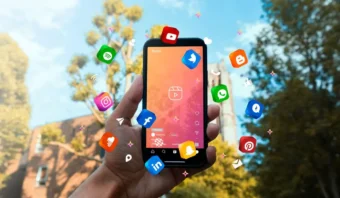 Una mano sostiene un teléfono inteligente al aire libre con varios íconos de aplicaciones flotando digitalmente alrededor del dispositivo, haciendo alusión a las mentiras de las redes sociales, todo ello con un telón de fondo de árboles y cielo azul.