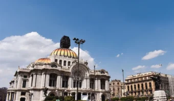 Palacio de Bellas Artes con una cúpula central y una fachada ornamentada bajo un cielo azul, rodeado de vegetación y edificios históricos en la vibrante capital de la Ciudad de México.