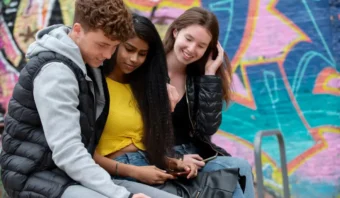 Tres jóvenes se sientan juntos al aire libre frente a una pared con grafitis de "Los Zetas", mirando un teléfono móvil y sonriendo.