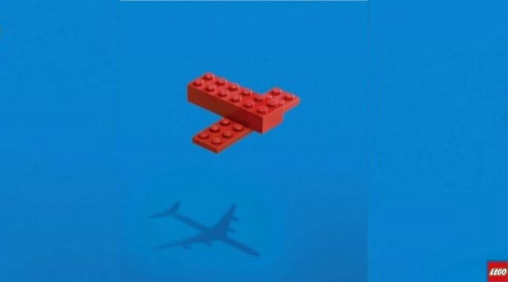 Dos ladrillos LEGO rojos forman la figura de un aviÃ³n simple sobre un fondo azul, y su sombra proyecta una imagen que recuerda a los aviones reales que suelen verse en los anuncios publicitarios.
