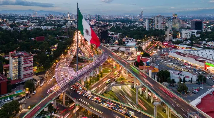 Vista aérea de un concurrido intercambio urbano en la Ciudad de México, la capital, con una gran bandera mexicana en el centro y edificios al fondo.