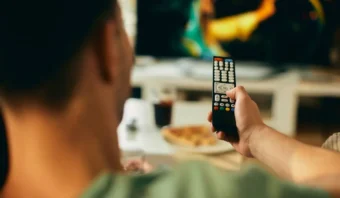 Una persona que sostiene un control remoto de TV lo apunta hacia un televisor, con una pantalla borrosa que muestra las calificaciones bajas, mientras que en el fondo se ven comida y bebidas.
