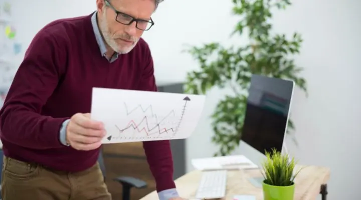 Un hombre que lleva gafas y un suéter marrón examina un cuadro gráfico impreso de los ratings de los niveles de los en un entorno de oficina con una computadora y una planta en el escritorio.