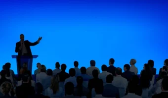 Un orador, despuÃ©s de tomar tiempo para preparar una conferencia, se sitÃºa en un podio y se dirige a una audiencia sentada en filas contra un fondo azul liso.