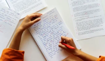 Una persona escribe con un bolígrafo rojo en un cuaderno, rodeada de páginas impresas llenas de notas manuscritas y correcciones centradas en la ortografía.