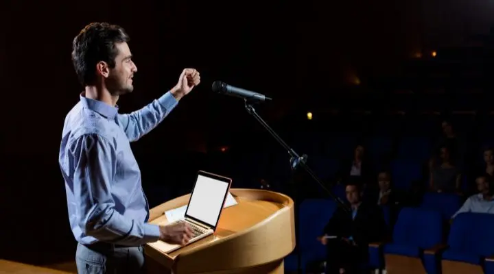Un hombre se encuentra en un podio con una computadora portátil, hablando por un micrófono a una audiencia en un auditorio con poca luz, mostrando cómo preparar una conferencia de manera efectiva.