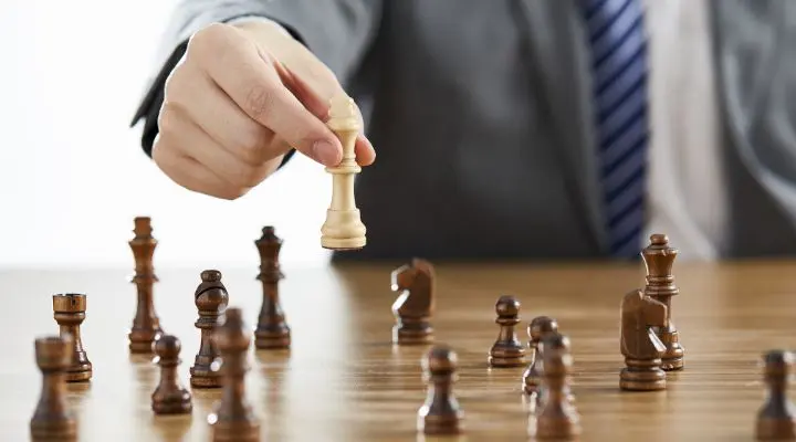Una persona con traje mueve pensativamente una pieza de ajedrez blanca sobre un tablero de madera durante un análisis de la partida.