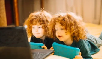 Dos niños pequeños, de pelo rojo y rizado, yacen en el suelo, sosteniendo tabletas azules y mirando la pantalla de una computadora portátil: una escena cotidiana que muestra la conexión entre los niños y la tecnología.