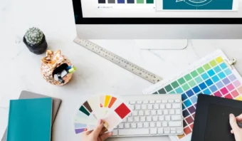 Una persona que sostiene muestras de color en un escritorio con un teclado, una tableta, una regla, muestras de pintura y un cactus decorativo usa espacio negativo en el diseño de logos mostrado en el monitor.