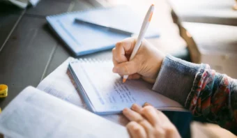Una persona escribe en un cuaderno con un bolígrafo, mientras que libros abiertos y otro cuaderno se encuentran sobre una mesa cercana, lo que refleja el enfoque en la imagen frente al texto durante el estudio.