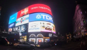 Grandes vallas electrónicas que muestran marcas importantes, como Coca-Cola, TDK y Hyundai, iluminan una calle concurrida llena de automóviles por la noche.