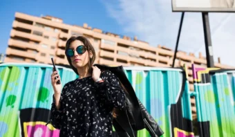 Una mujer que lleva gafas de sol y un vestido negro estampado sostiene un teléfono y una chaqueta mientras está parada frente a una valla colorida, con un edificio de apartamentos al fondo, capturando la energía vibrante que a menudo se ve en entornos de marketing callejero.
