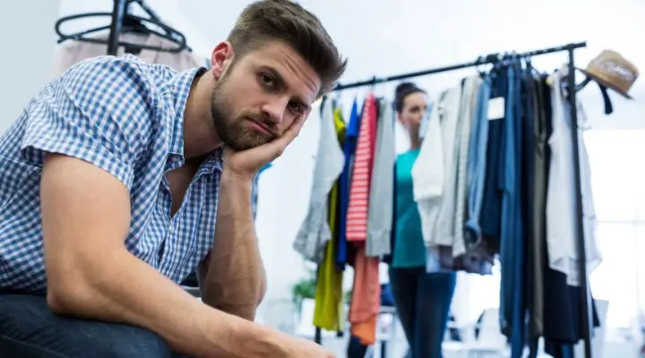 Un hombre se sienta con la cabeza apoyada en su mano, con aspecto aburrido, mientras una mujer compra ropa en una venta al fondo.