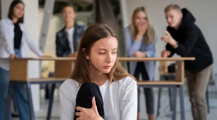 Una niÃ±a se sienta sola y parece triste mientras un grupo de adolescentes al fondo parecen chismorrear y seÃ±alarla, lo que pone de relieve el impacto del acoso laboral y la exclusiÃ³n social entre iguales.