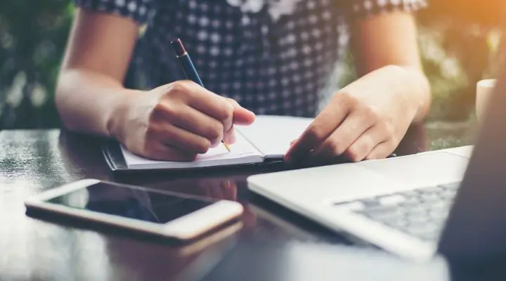 Una persona con una camisa a cuadros escribe en un cuaderno en una mesa con un teléfono inteligente y una computadora portátil, equilibrando imagen vs texto para tomar notas de manera efectiva.