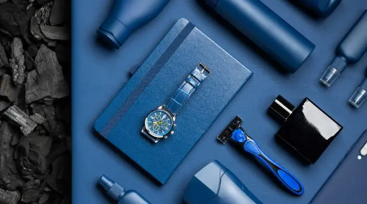 Plano de elementos de color azul, entre ellos un reloj, un cuaderno, una maquinilla de afeitar, botellas y un aerosol, dispuestos cuidadosamente sobre una superficie azul con carbón negro a la izquierda para contrastar.