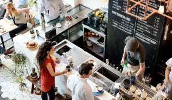 Una cafetería concurrida con personal preparando bebidas detrás del mostrador, el consumidor esperando y sentado, y un menú en la pizarra que enumera bebidas y pasteles.