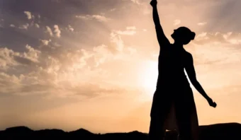 Silueta de una persona parada al aire libre con un brazo levantado contra un cielo al atardecer, representando una gran responsabilidad, con nubes dispersas y montañas en el fondo.