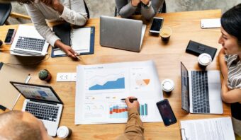 Cuatro personas se sientan alrededor de una mesa con computadoras portátiles, tazas de café, cuadernos y un gran cuadro comercial que muestra gráficos y datos, y discuten estrategias para un marketing de diálogo efectivo mientras una persona señala el cuadro.