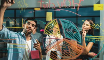 Cuatro personas realizan una lluvia de ideas sobre estrategias de marketing en una oficina, dibujando gráficos y diagramas en una pizarra transparente con marcadores de colores.