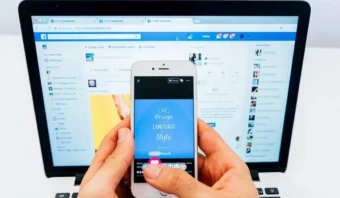 Una persona sostiene un teléfono inteligente que muestra una imagen con texto, frente a una pantalla de computadora portátil que muestra perfiles de redes sociales en una página de Facebook.
