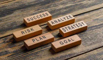 Bloques de madera sobre una superficie de madera rústica, cada uno etiquetado con una palabra relacionada con los negocios: ÉXITO, ANALIZAR, ESTRATEGIA, RIESGO, PLAN y OBJETIVO, lo que demuestra la consistencia retórica esencial para una comunicación empresarial eficaz.