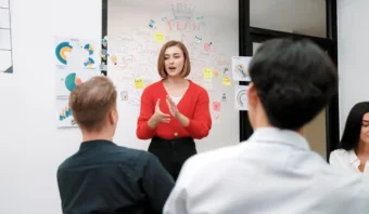 Una mujer con un cárdigan rojo se presenta ante un grupo en una oficina, abordando los desafíos que enfrentan los directores que no logran dirigir. Detrás de ella se ven gráficos y una pizarra con notas y diagramas.