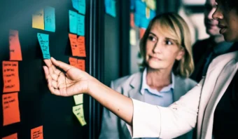 Tres personas estÃ¡n paradas frente a una pared cubierta con notas adhesivas de colores, intercambiando ideas mientras una extiende la mano para ajustar una nota: una escena interesante que genera nuevas perspectivas de marketing.