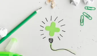 Un signo mÃ¡s verde con lÃ­neas garabateadas en forma de bombilla se encuentra sobre una superficie blanca, rodeado de un bolÃ­grafo verde, sujetapapeles, un marcador y papel arrugado: elementos que inspiran a los consumidores a despertar su creatividad.