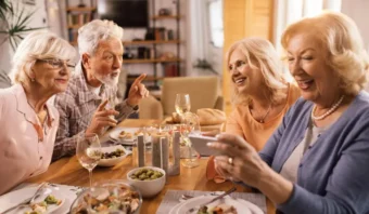 Cuatro adultos mayores, o seniors, se sientan alrededor de una mesa con comida y bebidas, sonriendo y hablando; una mujer mira su teléfono mientras los demás conversan.