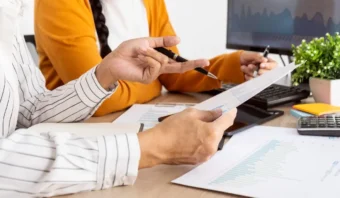 Dos personas se sientan en un escritorio revisando cuadros y gráficos impresos, con un monitor de computadora y suministros de oficina en el fondo, colaborando hacia una negociación exitosa.