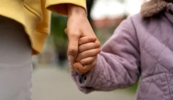 Primer plano de un adulto y un niño tomados de la mano al aire libre, simbolizando la conexión entre generaciones. El adulto lleva una manga amarilla; el niño, con una chaqueta acolchada lavanda, representa la nueva generación. El fondo está desenfocado.