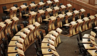 Filas de escritorios de madera vacíos y sillas negras dispuestas en un patrón curvo dentro de una moderna cámara legislativa o de conferencias, lo que refleja el potencial de reinvención política dentro de su cuidadoso diseño.