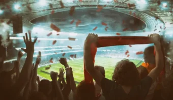 Una multitud de aficionados aplaude y levanta bufandas en un estadio, con confeti en el aire, las pancartas de la Asociación de Marcas ondeando orgullosamente y un campo de fútbol brillantemente iluminado en el fondo.