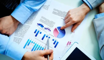 Dos personas vestidas de negocios revisan gráficos impresos y un gráfico circular en papel (quizás analizando datos como el informe de demografía maquillada del INEGI) con una tableta parcialmente visible sobre la mesa.