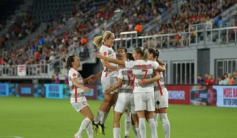 Un grupo de mujeres deportistas, vestidas con uniformes blancos, celebran juntas en el campo (una jugadora salta mientras las demás se abrazan), todo ello con el telón de fondo de una multitud en el estadio que las anima.