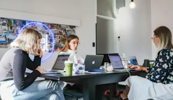 Tres mujeres se sientan en una mesa trabajando con computadoras portátiles en una oficina moderna y bien iluminada con un letrero de neón y fotos en la pared, perfecta para intercambiar ideas para ONs con tu agencia de publicidad.
