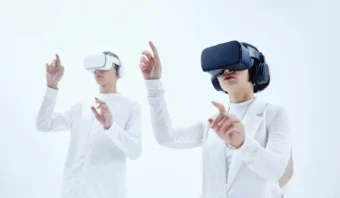 Dos personas con cascos de realidad virtual y ropa blanca se encuentran frente a un fondo blanco y levantan las manos como si estuvieran inmersos en un entorno de Realidad Virtual.