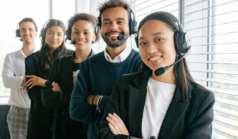 Cinco representantes de Servicio al Cliente con auriculares están de pie en fila en una oficina con grandes ventanales, sonriendo a la cámara.