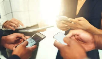 Cuatro personas paradas juntas en un área bien iluminada, sosteniendo y usando teléfonos inteligentes y una tableta, expresando sutilmente comunicación no verbal a través de sus gestos e interacciones.