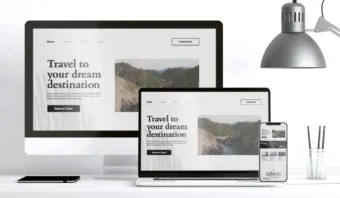 Un monitor de escritorio, una computadora portátil y un teléfono inteligente muestran el mismo sitio web de viajes, resaltando pensamiento responsivo en un escritorio blanco con una lámpara, una taza de lápices y un cuaderno cerrado.