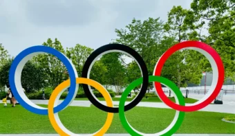 La escultura de los anillos olímpicos exhibida al aire libre en un parque, rodeada de césped verde y árboles bajo un cielo nublado, captura el espíritu compartido en las redes sociales en las olimpiadas.