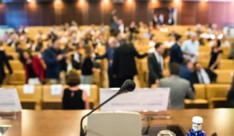 Un micrófono está ubicado sobre una mesa de conferencias en primer plano, con una audiencia borrosa, reunida para discutir sobre televisión y política, sentada y de pie en un auditorio en el fondo.